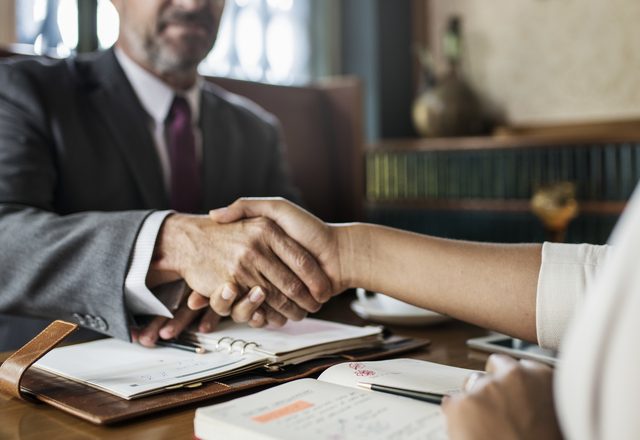Two people shaking hands across a desk during a business meeting